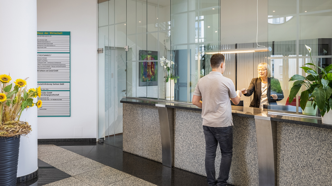 Das Bild zeigt eine Frau am Empfang, die einem Mann ein Dokument übergibt und dient als Titelbild für die Seite „Büro mieten in Dresden – Bürovermietung für Büroräume im Businesspark“.